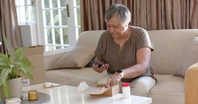 Mature African woman sitting on sofa opening shipping box and verifying bottle labels with phone