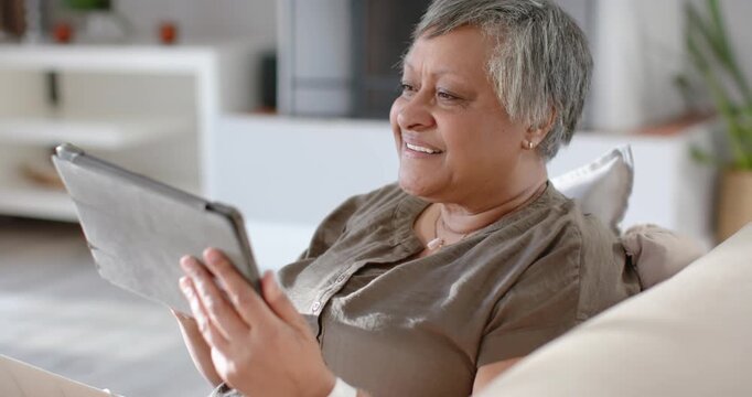 Senior African woman holding tablet and watching funny clip on sofa, adjusting grip, laughing