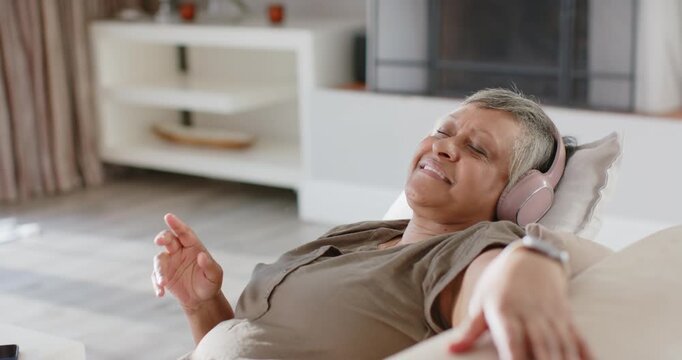 Senior African American woman reclining on sofa music playing on pink headphones tapping, settling