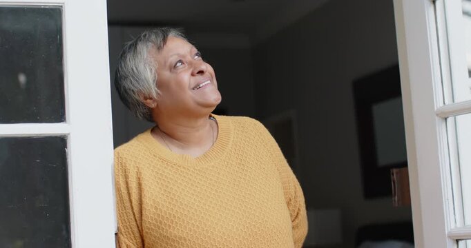 Senior African woman at glass door seeing outside motion, meeting camera, smiling in yellow sweater