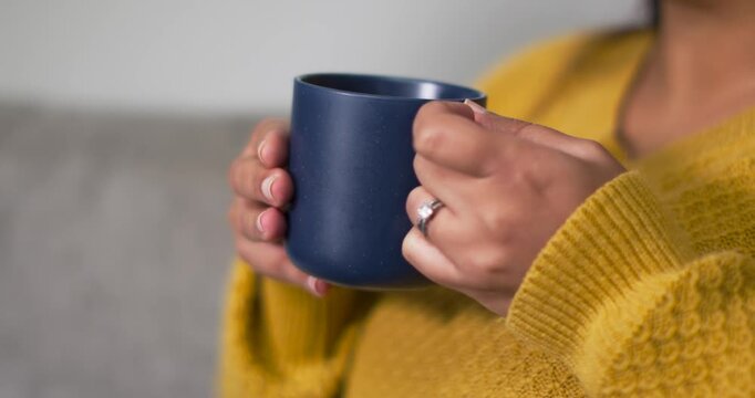 African American woman in mustard sweater cradling blue mug, lifting to sip on sofa, showing ring