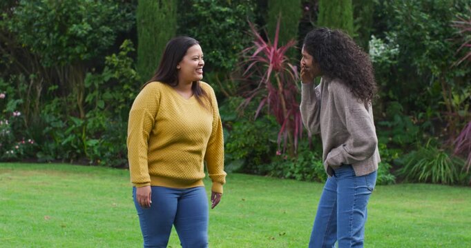 African American female friends standing by shrubs on lawn, right woman starting story, gesturing