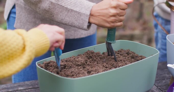 African American women breaking packed soil in green planter with trowel and fork, planting seeds