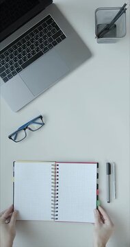 Hands placing open spiral notebook with checkered paper on white table near laptop, glasses, pen, pencil, and pencil holder, ideal for study, work, or creative projects.