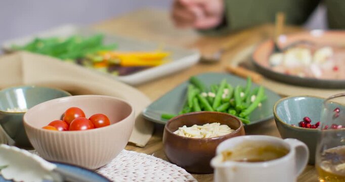 As camera racking African-American woman in sweater reaching spoon to serve bowls on dining table