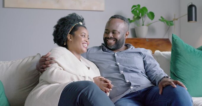 Adult African couple sitting on couch at home with pillows, woman speaking, man listening, laughing