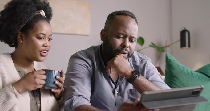 African American couple on sofa woman holding blue mug pointing tablet guiding man tapping smiling