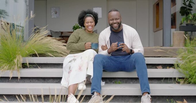 Couple exchanging smiles sitting on front steps holding blue mugs, leaning and sharing warmth