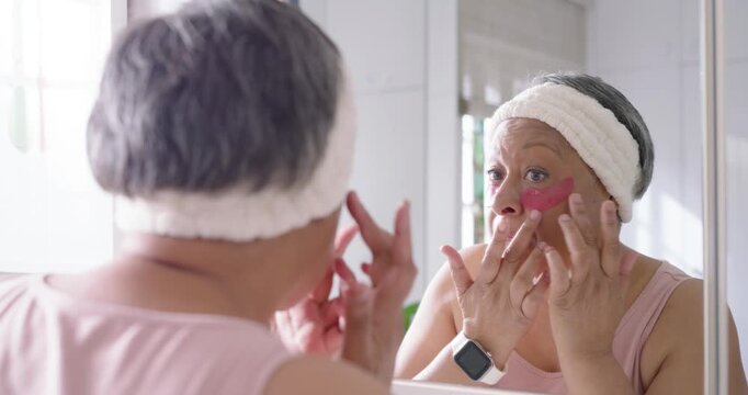 Senior African American woman looking into mirror applying pink eye patches for skincare at sink