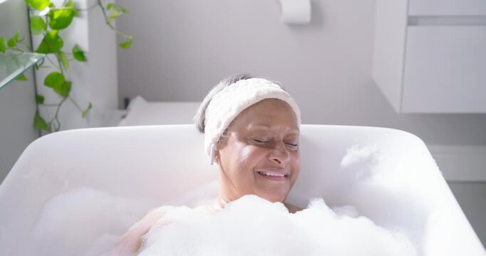 Senior African American woman soaking in bathroom tub with headband foam for calm smiling gesturing