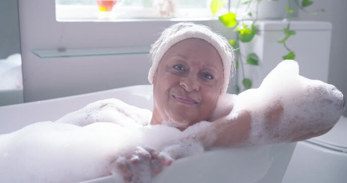 Senior African American woman lying in tub rubbing foam opening eyes smiling as light drawing gaze