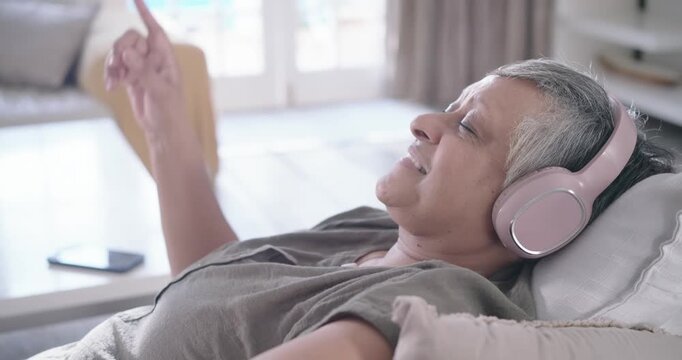 Senior African woman lounging on sofa, wearing headset, nodding, gesturing to cell music, settling