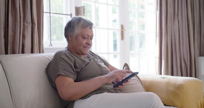 African mature woman examining card picking phone checking message smiling on sofa by French doors