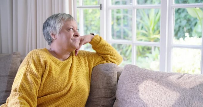 Senior African American woman sitting on sofa by window, sun drawing gaze, smiling in yellow knit