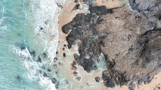 Aerial view of turquoise waves crashing against the dark jagged rocks and sandy shore of Constantine Bay in Padstow, Cornwall, United Kingdom.