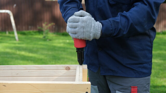 Close-up of worker in blue uniform and safety gloves using a red pneumatic nail gun to assemble a wooden structure outdoor. Green grass blurred background. Home improvement and DIY concept. Banner