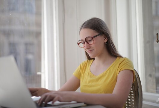 Young woman with glasses works on laptop by window