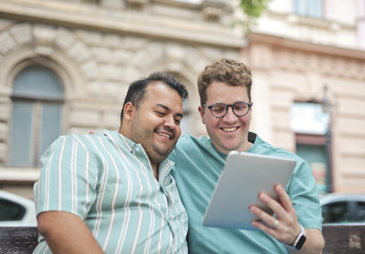 Happy male couple sharing a tablet on a city bench