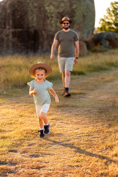 Happy little girl running ahead of dad on walk in rural paddock reserve wearing Akubra hat
