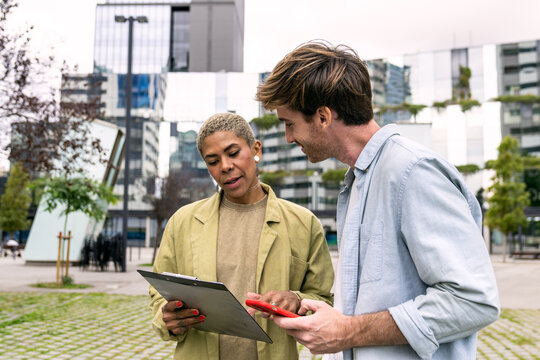 Business people collaborating and communicating outdoors, reviewing documents on a clipboard and interacting with a red smartphone