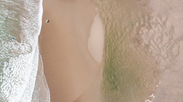 Aerial view of surfers walking on the sandy shore of Bantham Beach with crashing waves and textured sand ripples in Kingsbridge, Devon, United Kingdom.