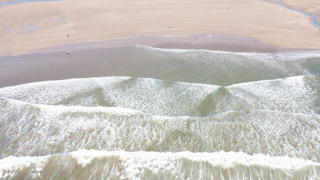 Aerial view of Bantham Beach with rolling waves and white foam on the sandy shore as surfers walk along the water in Kingsbridge, Devon, United Kingdom.