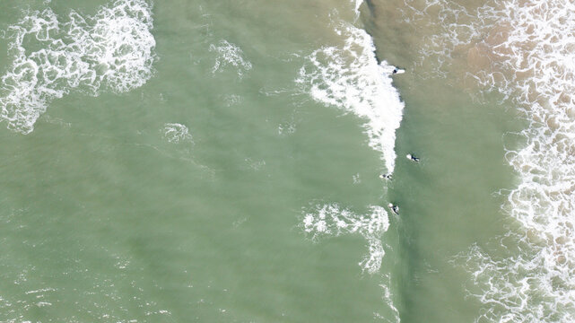 Aerial view of surfers waiting for waves in the turquoise ocean with white foam from breaking waves at Bantham Beach, Kingsbridge, Devon, United Kingdom.