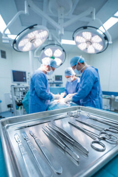 Close-up of sterile stainless steel surgical instruments in a tray. Blurred medical team of surgeons performing an operation under bright lights in a hospital operating theater.