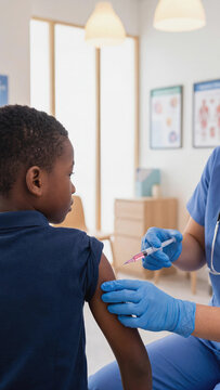 A young Black boy getting a vaccine shot in his arm from a healthcare worker at a clinic. Female doctor in blue scrubs and gloves administering a liquid medicine injection from a syringe.