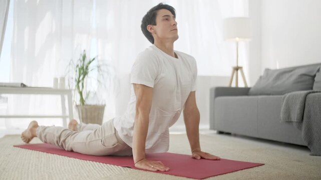Focused man performing upward facing dog yoga pose on a mat in a bright modern apartment. Young male, person or adult stretching his chest, shoulders and back during a home workout, wellness practice