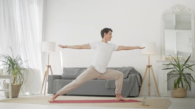 Athletic man practicing a high lunge yoga pose on a mat in a spacious modern apartment. Young male, person or adult exercising barefoot with raised arms, following a smartphone class, online training