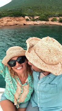 Joyful middle-aged couple wearing straw hats taking a selfie video while sailing on a boat, smiling and kissing, with a beautiful green coastline in the background during a summer vacation