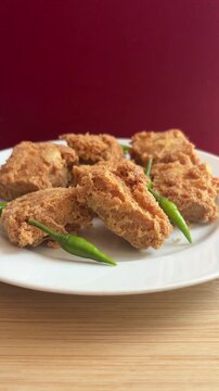 Crispy fried tofu or Tahu Crispy in cube pieces on a white plate. Small green chilies add spice. Red backdrop contrasts with warm food tones. Wooden surface beneath the plate. indonesian street food