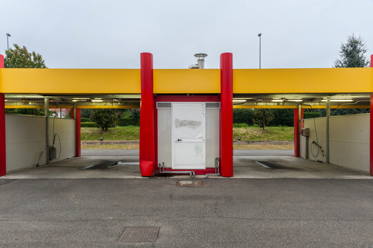 Architettura minimalista di una stazione di servizio con colonne rosse audaci e una pensilina gialla. La struttura &egrave; circondata da una superficie in asfalto grigio e da un edificio in pietra.