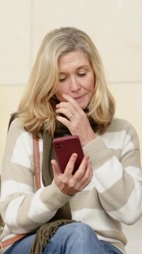 Vertical video: Looking down, woman holding red phone and scrolling on bench, adjusting scarf