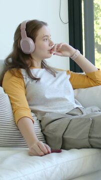 Vertical video: Sitting woman in mustard top holding phone on couch at window, light drawing gaze