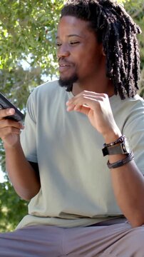 Vertical video: Smiling man in green tee sitting on bench under tree reading phone and laughing