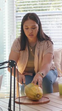 Vertical video: Placing papaya on board, woman slicing with chef knife at window, preparing to eat