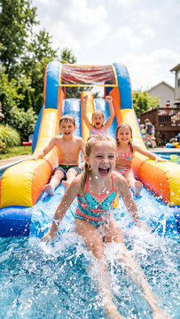 Four excited Caucasian children sliding down a colorful backyard inflatable water slide on a hot summer day. Happy boys and girls splashing into a pool during a fun outdoor party.