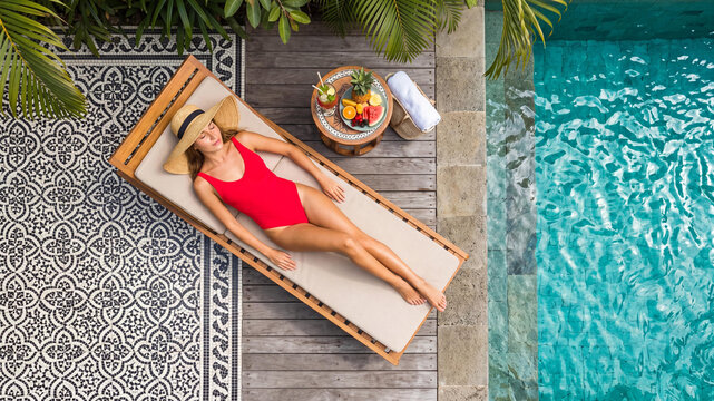 Overhead top view of a young Caucasian woman in a red swimsuit and straw hat relaxing on a wooden lounger by a turquoise pool. Luxury tropical resort villa backyard on a sunny summer day.