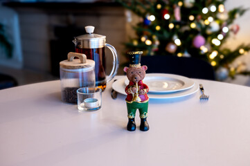 A festive decorative bear figurine in a suit stands beside a glass French press, a jar and white plates on a white table before a glowing Christmas tree. A cozy holiday table scene with copy space. © Oksana