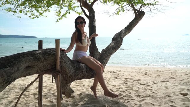Happy woman on sand beach, sitting on a large rustic tree branch with her arms wide open, wearing a white bikini top and sunglasses, experiencing ultimate freedom and joy during a sunny summer vacatio