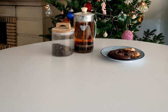 A glass French press of tea, a tea jar and a plate of chocolate cookies on a white table before a glowing Christmas tree with a fireplace. A cozy minimal holiday drink scene with warm lights and copy