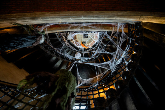 White cobwebs drape a dark vintage staircase with hanging decorations creating a haunted Halloween atmosphere. An eerie indoor scene of festive Halloween decoration in an old building.