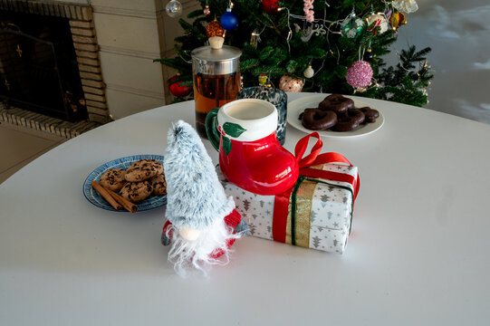 A Christmas gnome, a red Santa boot mug on a wrapped gift, a French press of tea and plates of cookies on a white table before a glowing Christmas tree. A cozy holiday treats and drink scene with copy