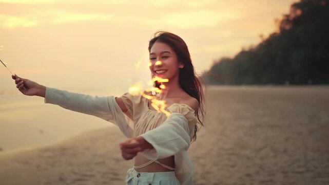 Happy Asian woman holding sparklers and smiling on beach at sunset, cheerful young female enjoying summer vacation and celebrating on seashore during golden hour for travel, holiday, and freedom.