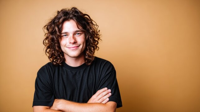 A young man with long curly hair confidently poses with arms crossed, exuding charisma and self-assurance, set against a warm, solid color backdrop.