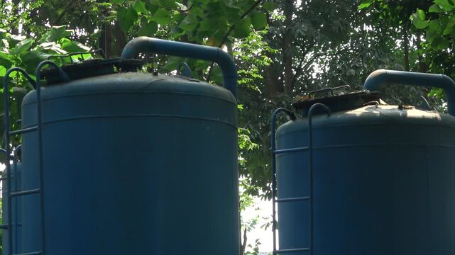 Low angle camera movement tracking the top section of large blue water reservoir cylinders and connection pipes, framed against dense green tree leaves and tropical foliage in a public urban park.