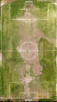 Zenithal aerial view of an abandoned, ruined soccer field with faded white lines, featuring worn patches of bare dirt and wild, patchy green grass growing unevenly across the entire pitch.