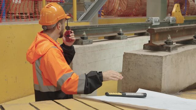 Serious construction worker intently studies detailed bridge blueprint amidst active building site operations, exhibiting focused engineering work and meticulous planning near wooden surface.